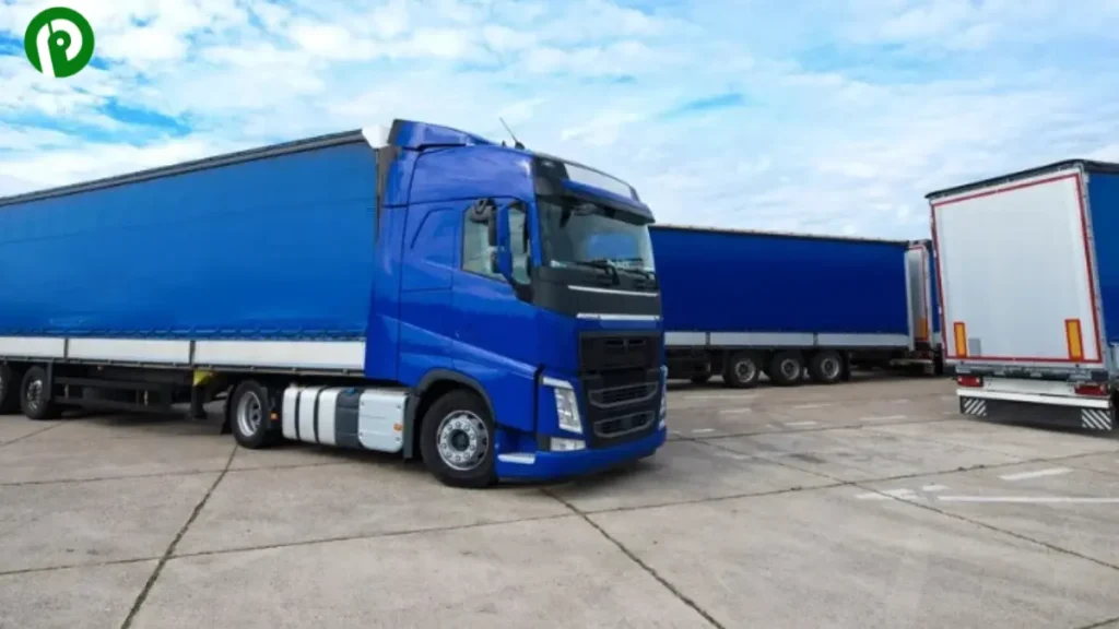 Two blue semi-trailer trucks parked on a concrete lot with a partly cloudy sky in the background.