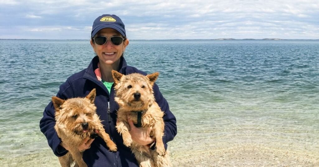 Jill Schlesinger standing by a scenic ocean shore, smiling and holding two small wet terrier dogs, wearing a navy jacket, sunglasses, and a baseball cap.