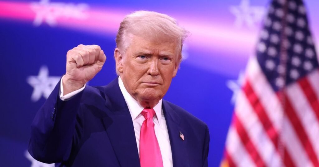 Donald Trump raising his fist at a political event with American flag in background.
