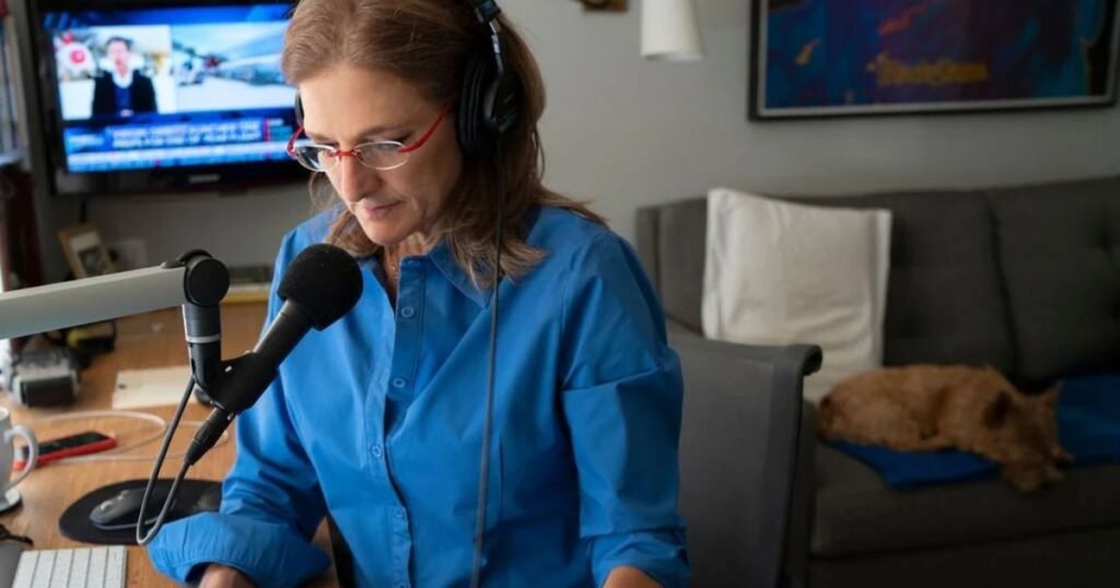 Jill Schlesinger wearing a blue shirt and red glasses, speaking into a microphone in her home studio setup, with a small terrier dog resting on a couch behind her.