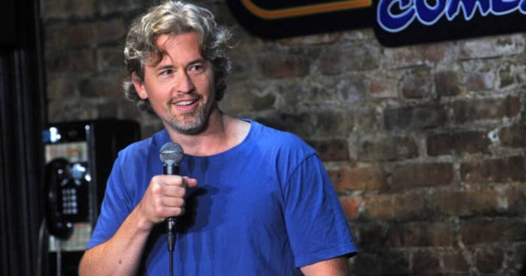Matt McCusker performing stand-up comedy in a blue shirt, holding a microphone.