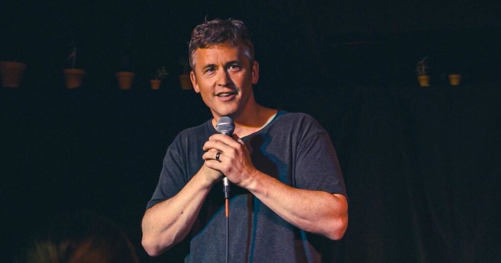 Matt McCusker performing stand-up comedy on stage, holding a microphone.