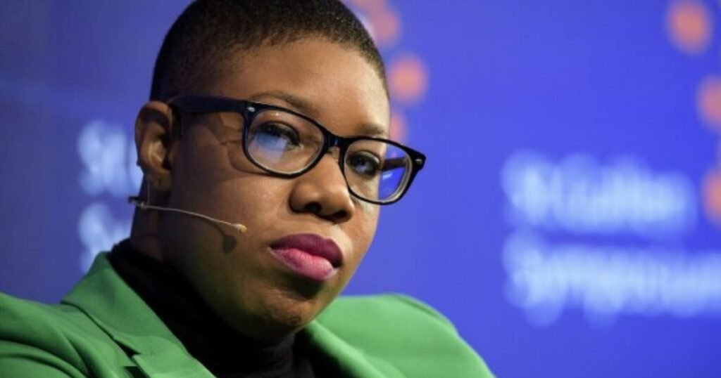 Symone Sanders wearing glasses and a green blazer while speaking at a professional event with a microphone headset.