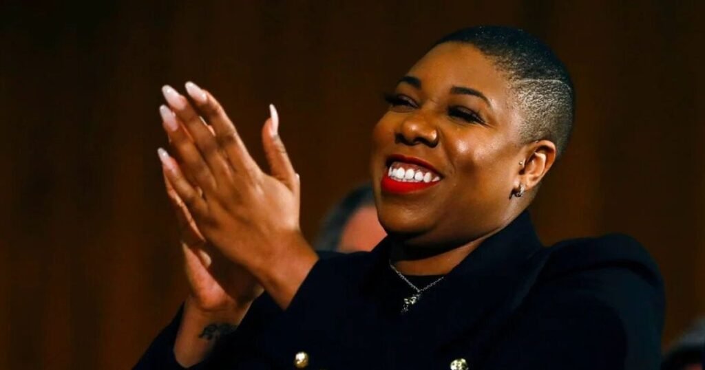 Symone Sanders smiling and clapping, wearing a black outfit and red lipstick.