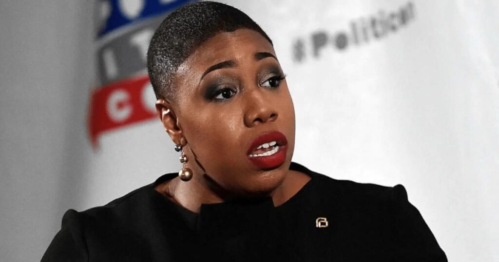 A close-up of Symone Sanders speaking passionately at a public event, wearing a black outfit and pearl earrings.